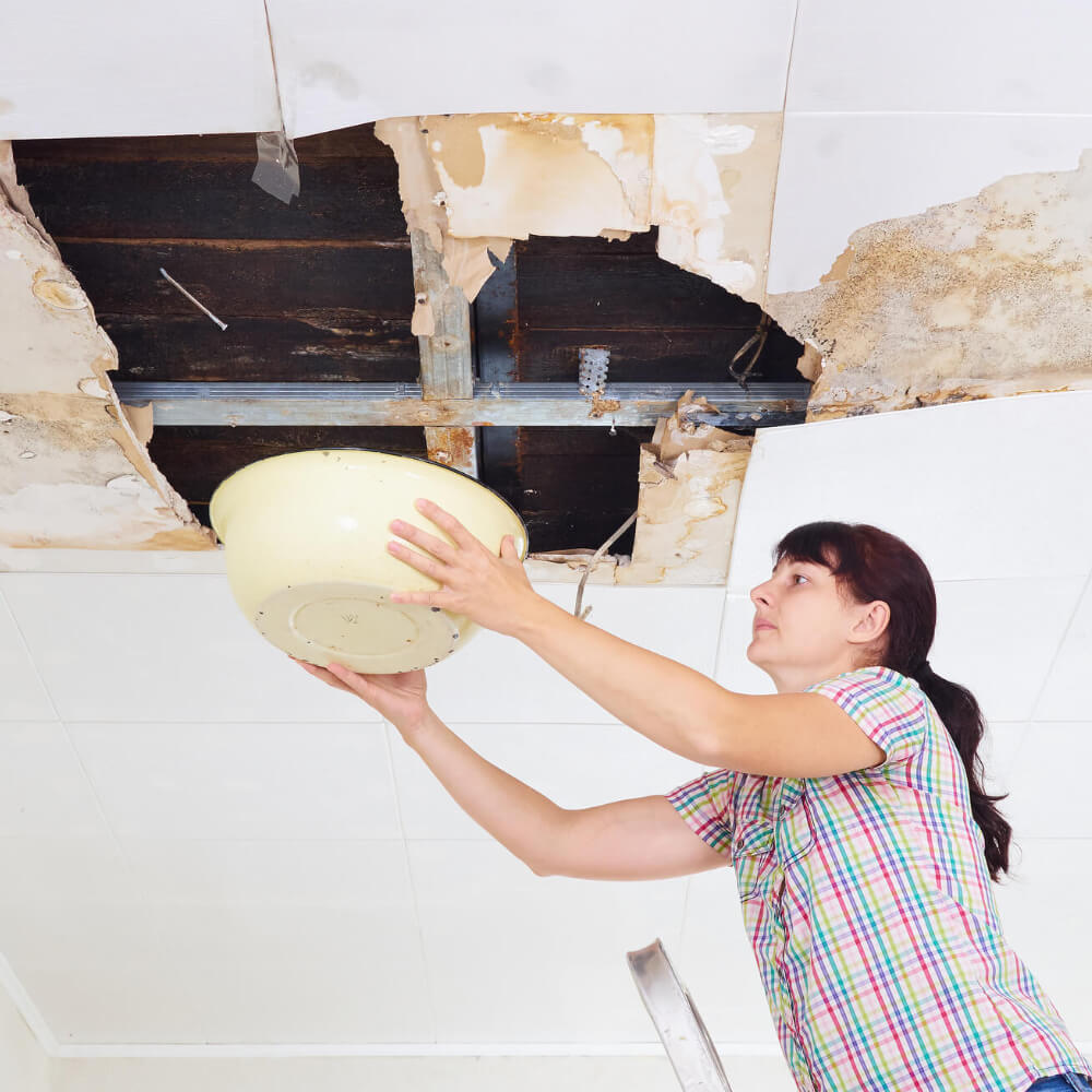 Picture of a woman using a bucket to catch water due to roof's leaking.